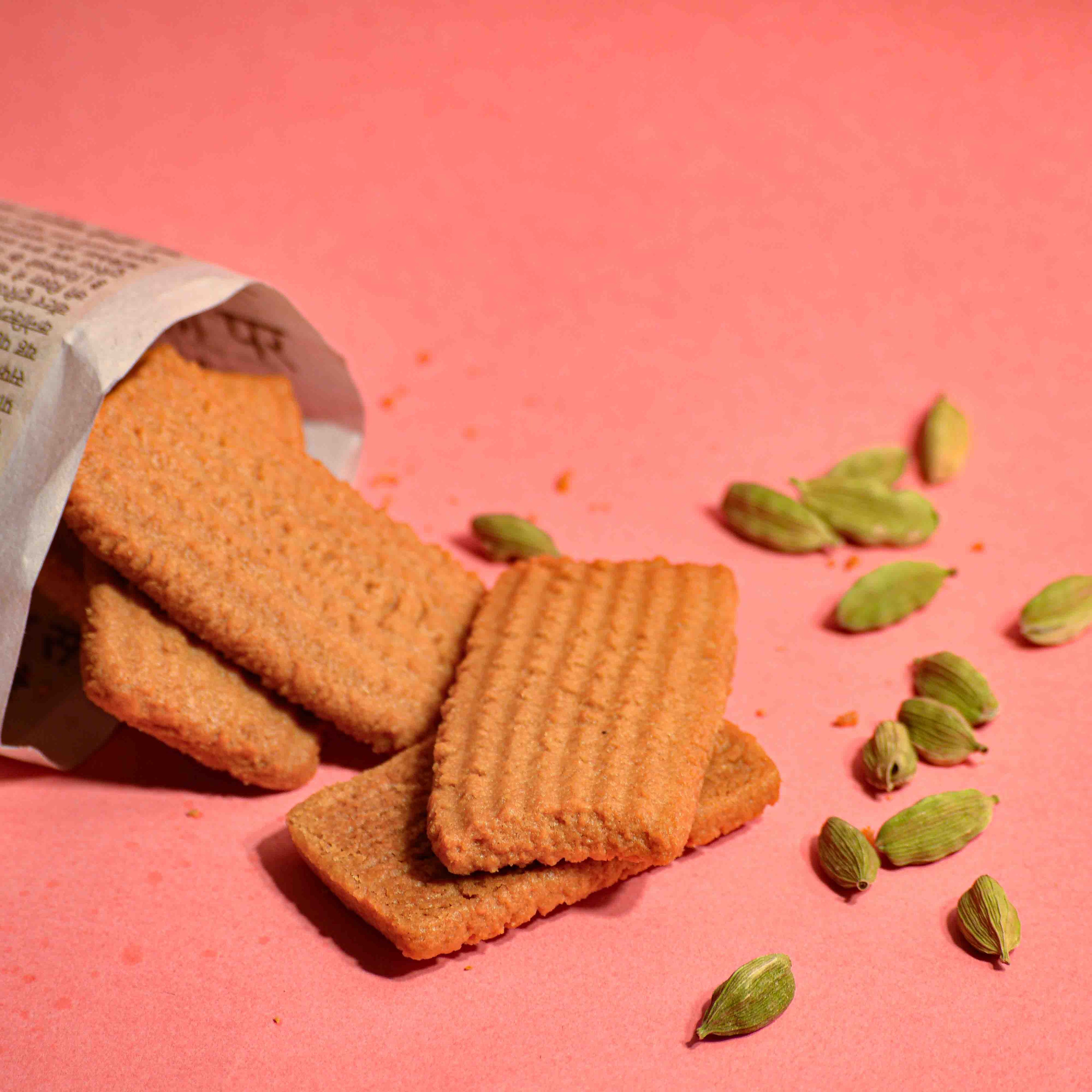 Indian biscuit atta biscuit stacked on a pink background with cardamon pods scattered around professional product photography