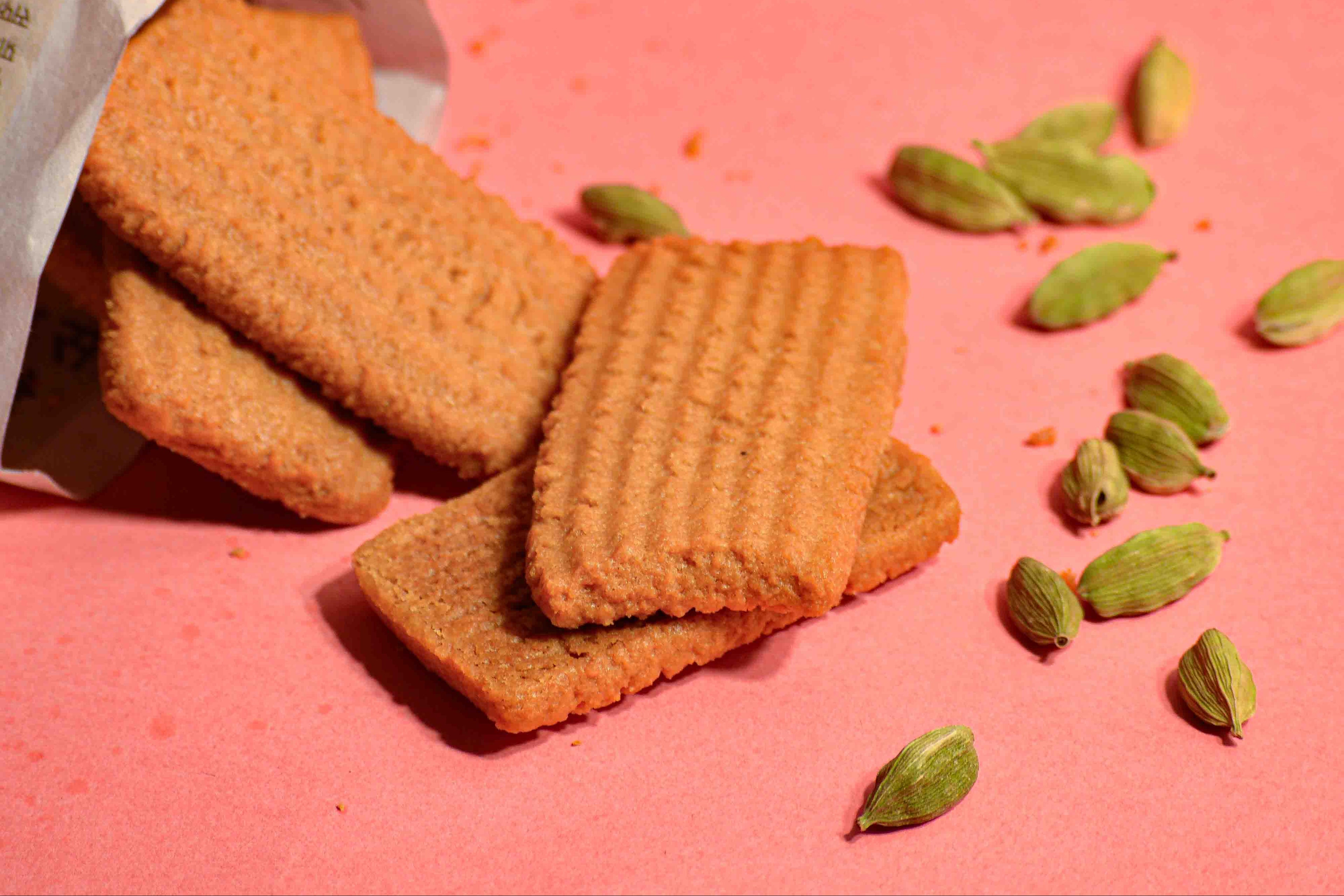 Indian biscuit atta biscuit stacked on a pink background with cardamon pods scattered around professional product photography