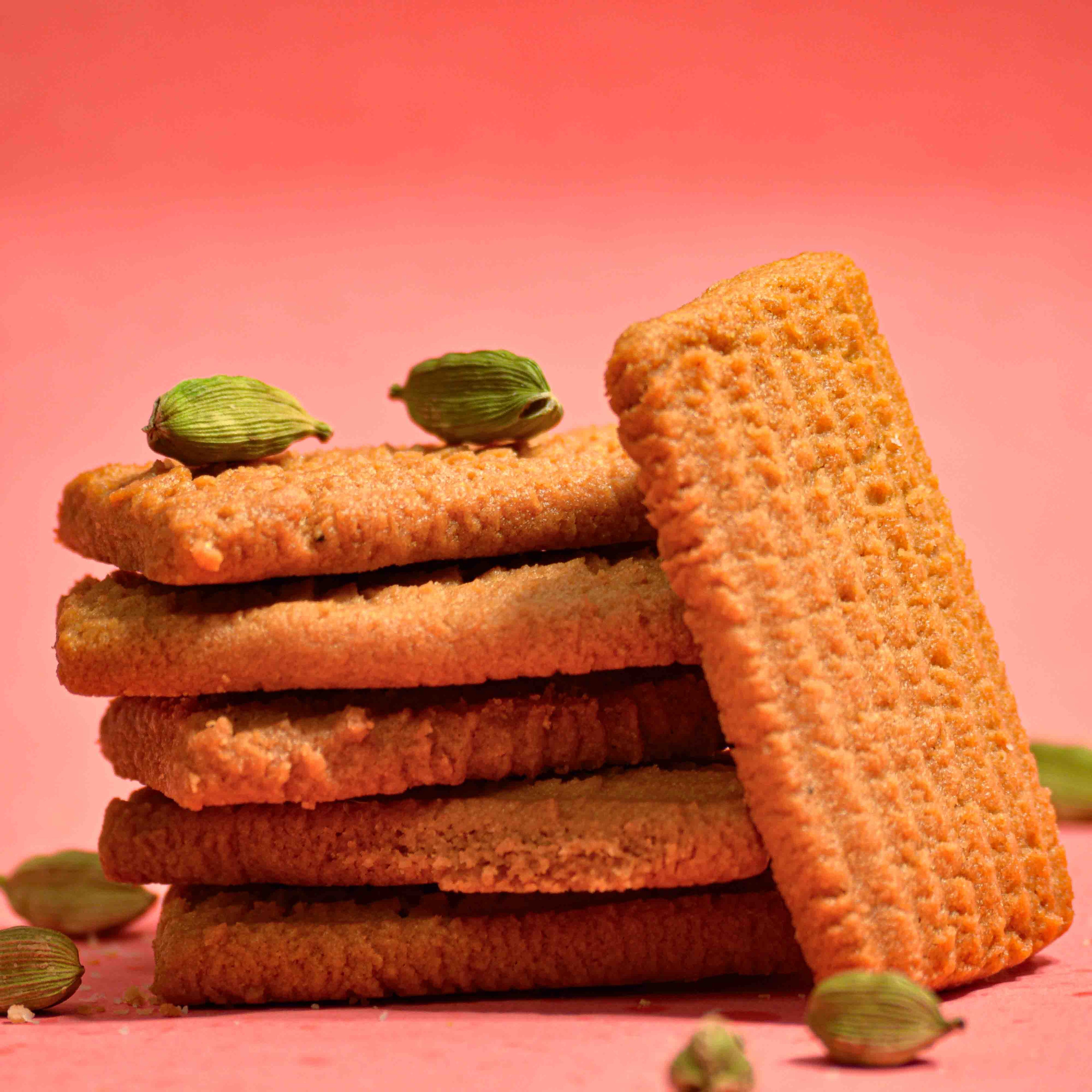 Indian biscuit atta biscuit stacked on top of each other with pink background with cardamon pods professional product photography